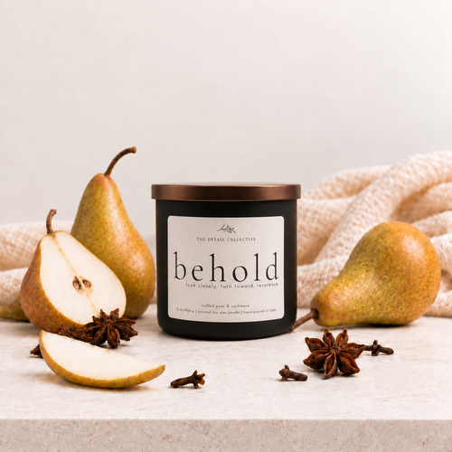 black candle titled 'behold' with a cashmere blanket in the background, pears and spices around it.