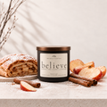 A candle jar labeled 'believe' by The Artful Collective, styled with baked apple strudel, apple slices, cinnamon sticks, and powdered sugar on a light stone surface against a warm neutral background.