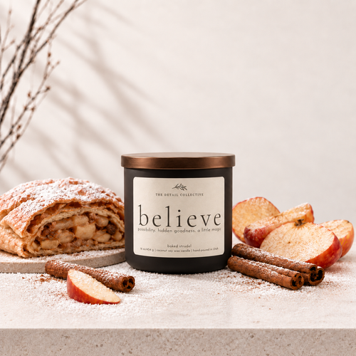 A candle jar labeled 'believe' by The Artful Collective, styled with baked apple strudel, apple slices, cinnamon sticks, and powdered sugar on a light stone surface against a warm neutral background.