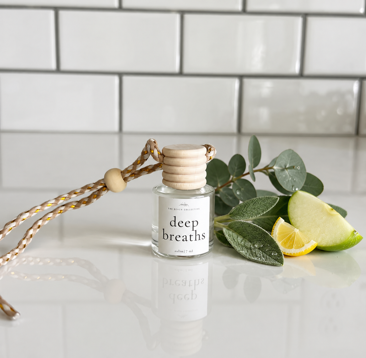 A glass reed diffuser bottle labeled 'deep breaths' with a light wood cap and braided cord, styled on a glossy white surface with eucalyptus leaves, a lemon slice, and a lime wedge against a white subway tile background.