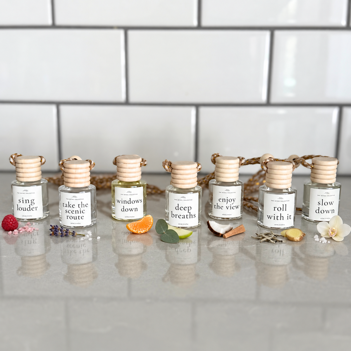 7 car diffusers lined up on a cool stone surface labeled 'sing louder,' 'take the scenic route,' 'windows down,' 'deep breaths,' 'enjoy the view, 'roll with it,' and 'slow down'. there are scent images surrounding each bottle, fruit, sugar flowers, orange slices, greenery, apple, coconut, wood, tea, and sea salt