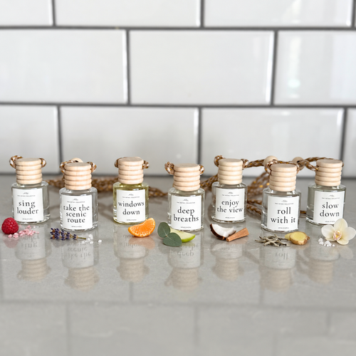 7 car diffusers lined up on a cool stone surface labeled 'sing louder,' 'take the scenic route,' 'windows down,' 'deep breaths,' 'enjoy the view, 'roll with it,' and 'slow down'. there are scent images surrounding each bottle, fruit, sugar flowers, orange slices, greenery, apple, coconut, wood, tea, and sea salt