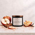 A black glass candle jar with a metallic copper lid and a white label reading 'gather' by The Royal Collection, styled on a light stone surface with sliced red apples, cinnamon sticks, and vanilla beans against a soft light gray background.