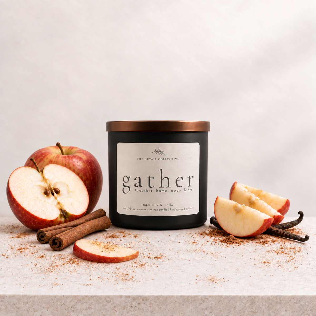 A black glass candle jar with a metallic copper lid and a white label reading 'gather' by The Royal Collection, styled on a light stone surface with sliced red apples, cinnamon sticks, and vanilla beans against a soft light gray background.