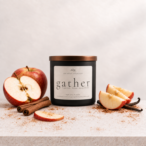 A black glass candle jar with a metallic copper lid and a white label reading 'gather' by The Royal Collection, styled on a light stone surface with sliced red apples, cinnamon sticks, and vanilla beans against a soft light gray background.