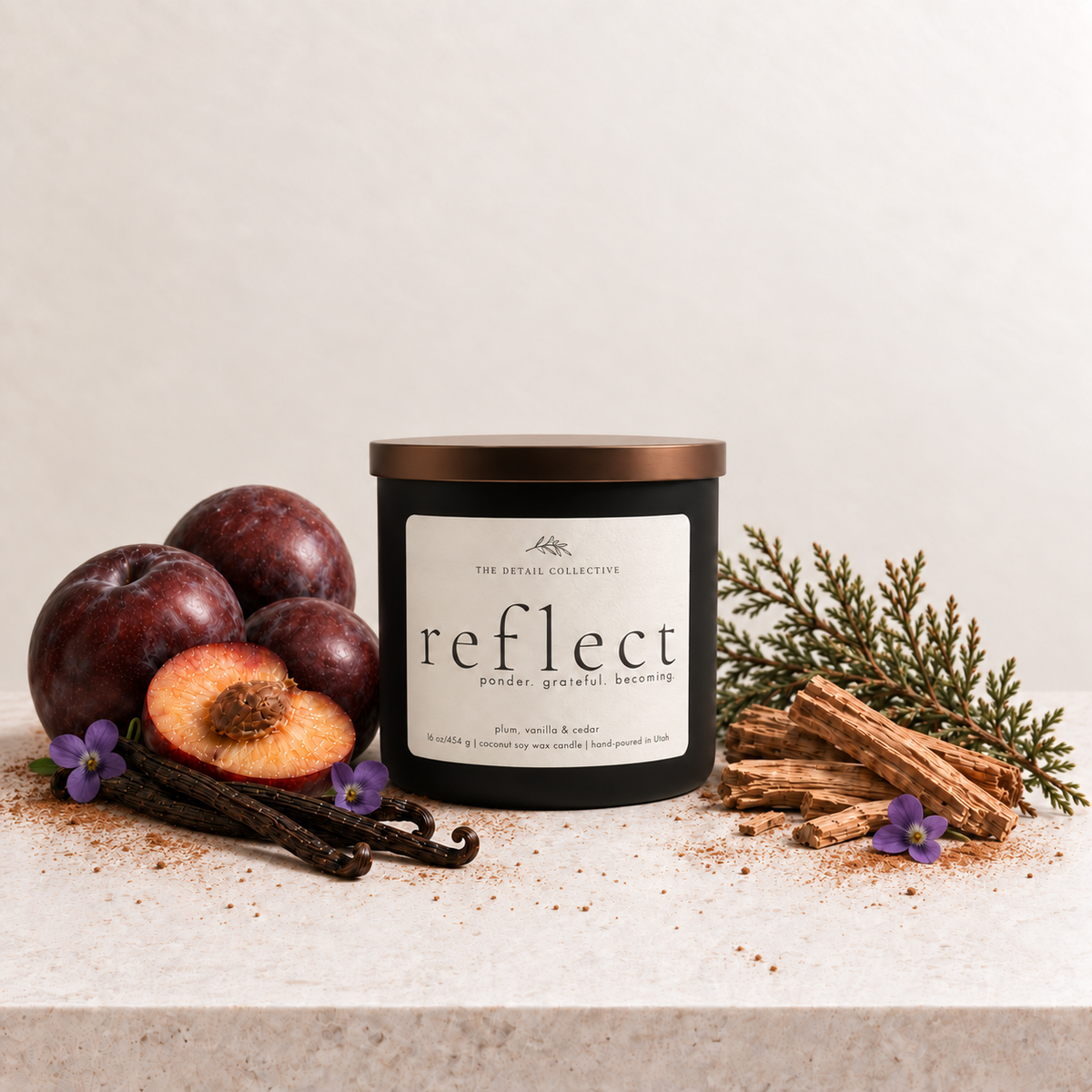 A matte black candle container with a bronze  lid and an ivory  label reading 'reflect' by the 'The Detail Collective' sits on a stone surface with plums, vanilla beans, and cinnamon sticks around it.