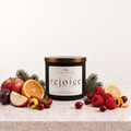 A matte black candle container with bronze lid from The Detail Collective labeled 'rejoice' on an ivory label, sits on a light surface with fruit props including plums, raspberries, sliced fruit, and a cinnamon stick against a warm neutral background.