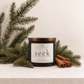 A matte black candle container with a bronze lid and ivory label reading 'seek' by The Detail Collective, styled on a stone surface with pine branches and cinnamon sticks.