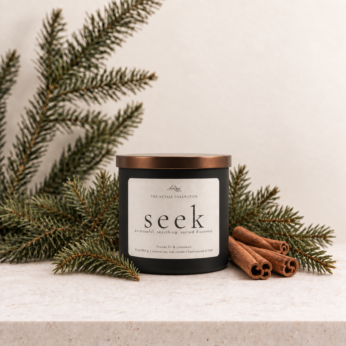 A matte black candle container with a bronze lid and ivory label reading 'seek' by The Detail Collective, styled on a stone surface with pine branches and cinnamon sticks.