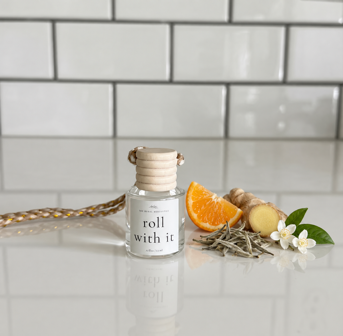 A glass diffuser bottle labeled 'roll with it' sits on a glossy white surface with a tiled backsplash behind it. The bottle is styled with a braided cord, an orange slice, ginger root, dried herbs, and small white flowers.