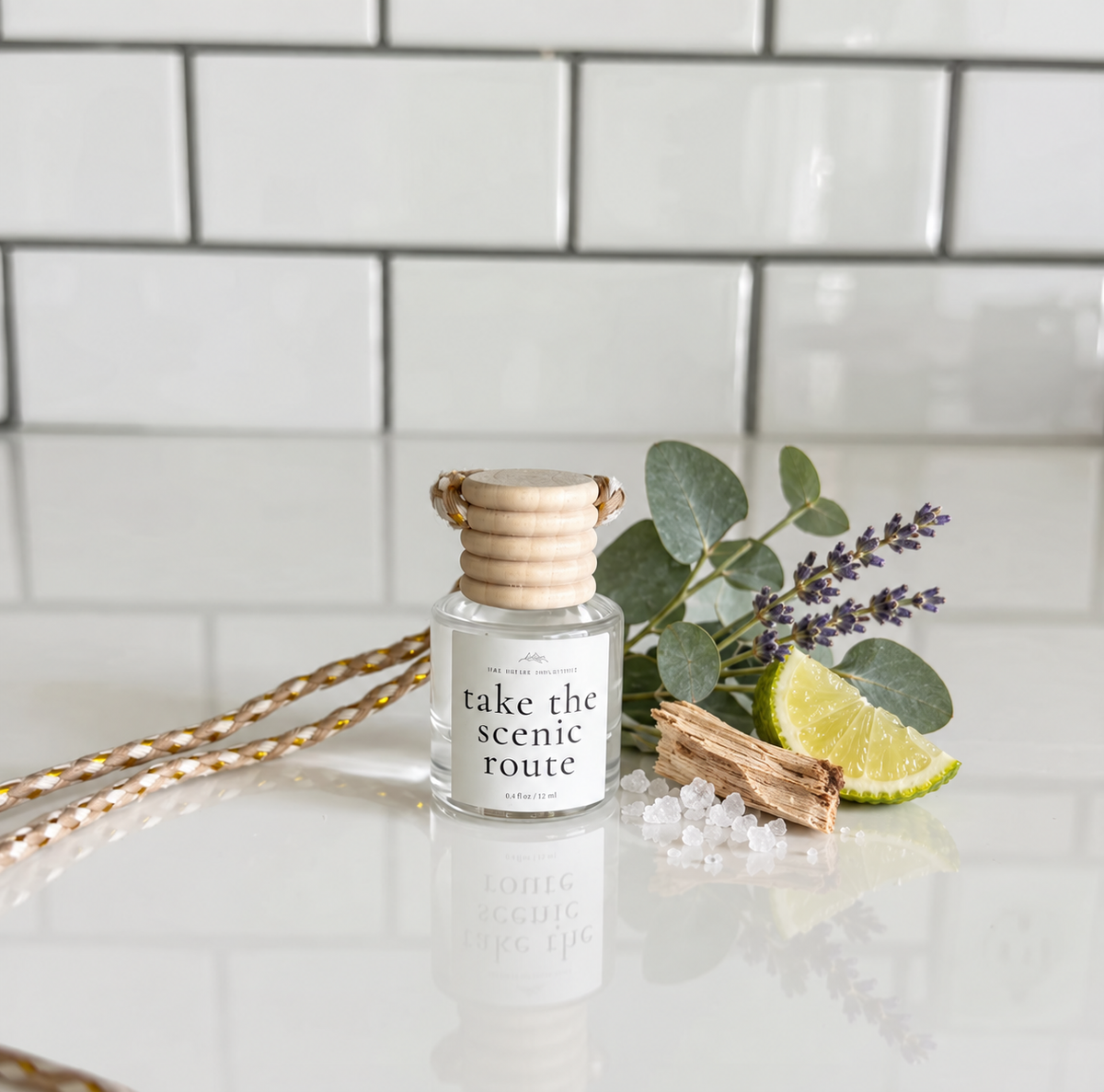 A small glass reed diffuser bottle labeled 'take the scenic route' sits on a glossy white surface with decorative eucalyptus leaves, lavender sprigs, a lime slice, a cinnamon stick, and a gold-and-white braided cord against a white subway-tile background.