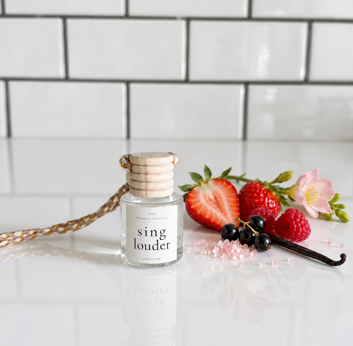 A small glass reed diffuser bottle labeled 'sing louder' sits on a glossy white surface with a woven reed collar and hanging cord. The bottle is styled with fresh strawberries, raspberries, blackberries, a vanilla bean, and pink salt crystals against a white subway-tile background.