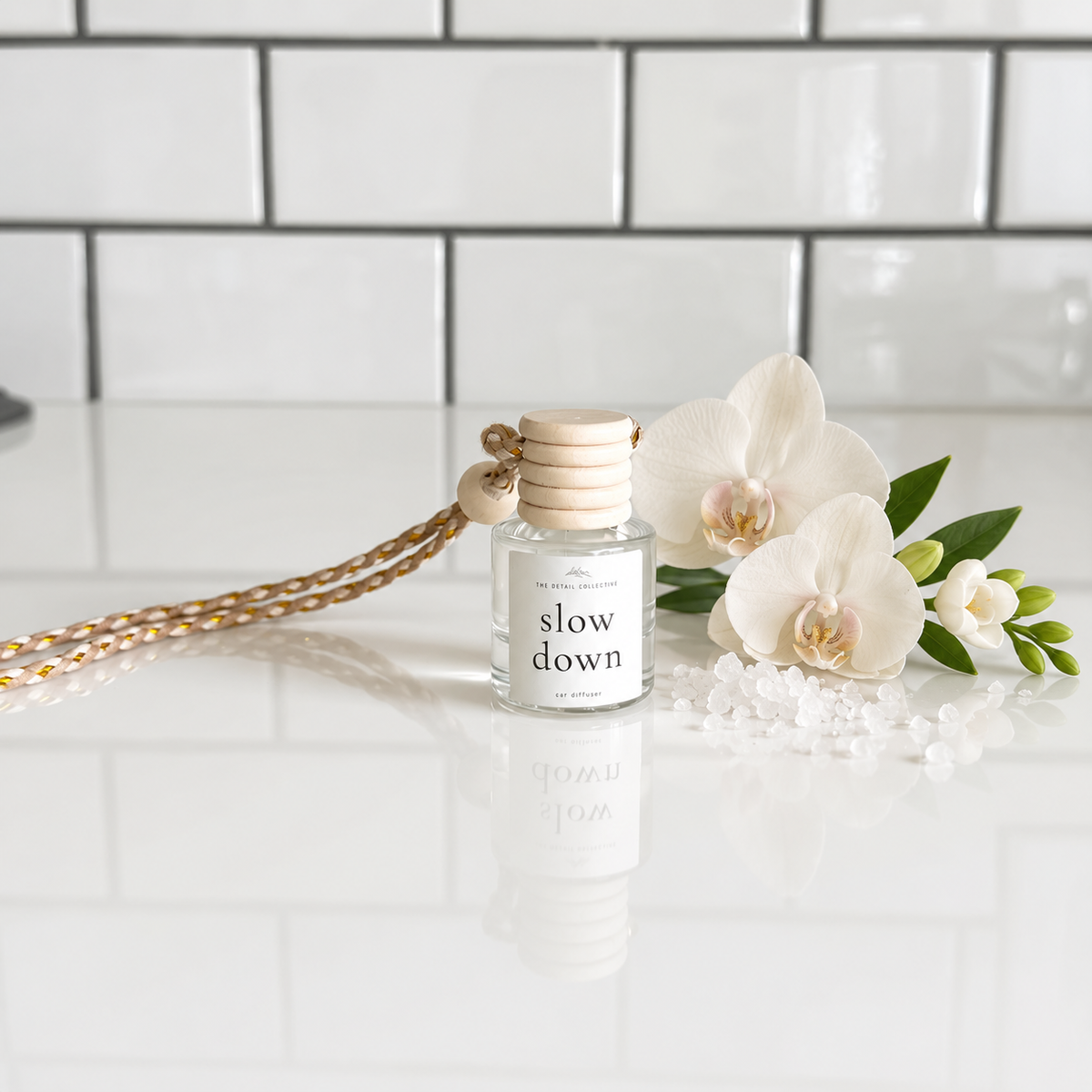 A clear glass reed diffuser bottle with a light wood cap and a gold-toned hanging cord, labeled 'slow down' and styled on a glossy white surface with white flower petals and decorative beads against a white subway tile background.