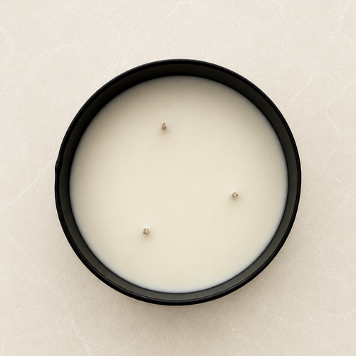 A top-down view of a black round candle container filled with white wax, featuring three small wicks, set against a light beige stone-textured background.