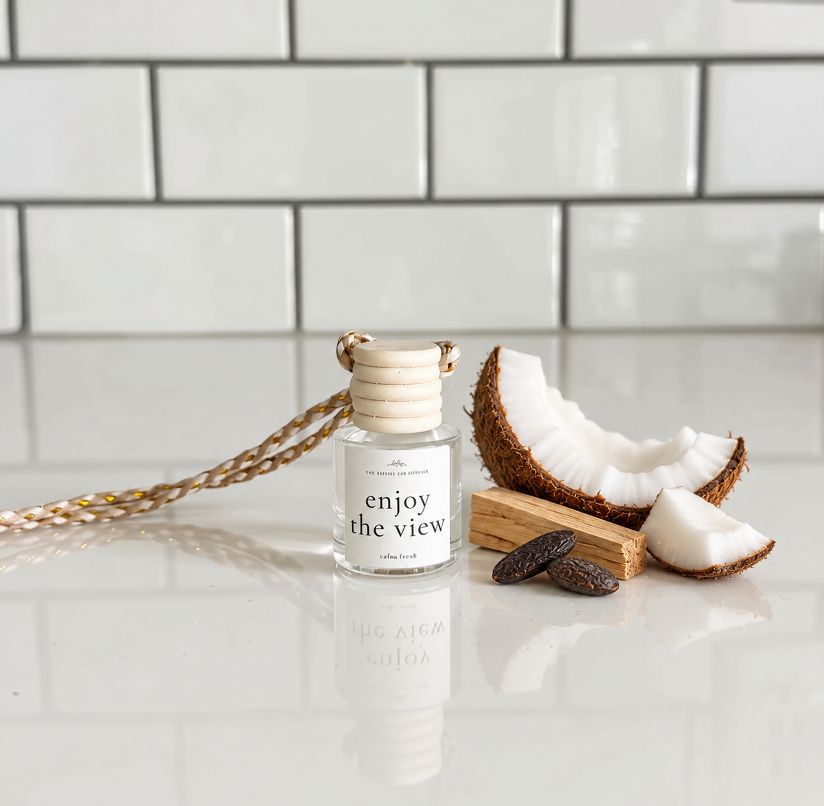 A glass diffuser bottle labeled 'enjoy the view' sits on a glossy white surface with a tiled backsplash behind it. The bottle is styled with a braided cord, pieces of coconut, sandalwood and tonic beans.