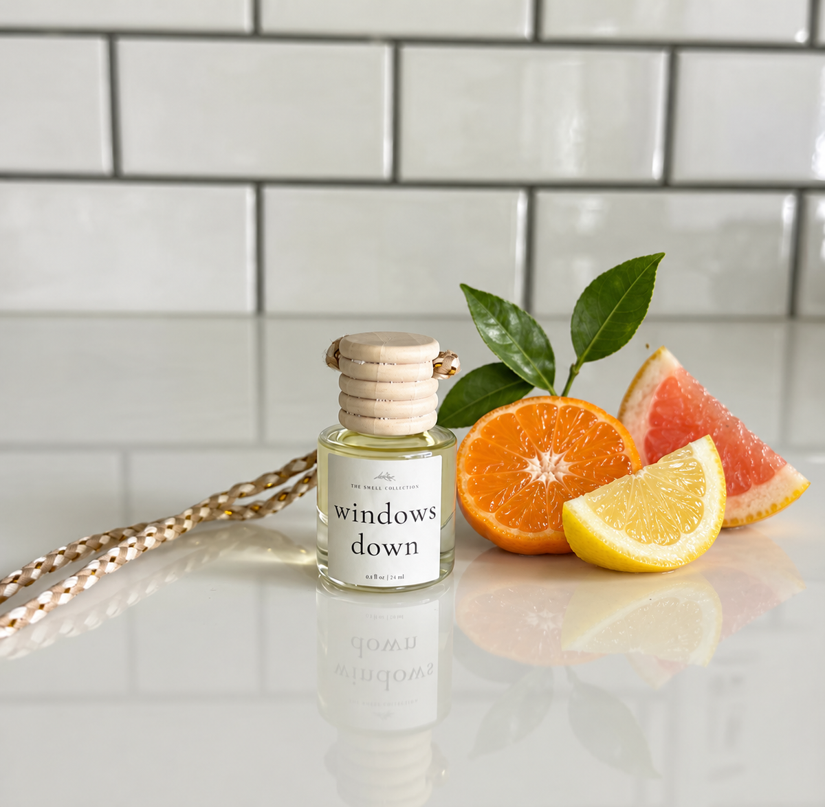 A reed diffuser bottle labeled 'windows down' sits on a glossy white surface with citrus slices and green leaves beside it, against a white subway-tile background.